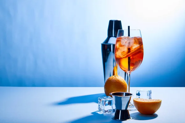 Cóctel Aperol Spritz, naranjas, coctelera, cubitos de hielo y taza de medir sobre fondo azul - foto de stock