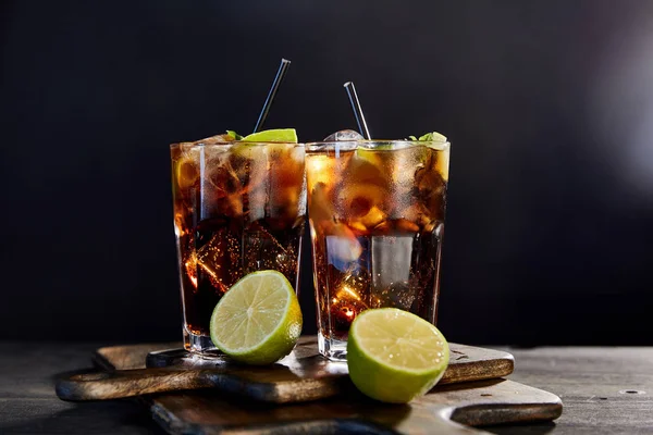 Coquetéis cuba libre em copos com palhas e limas sobre fundo preto — Fotografia de Stock