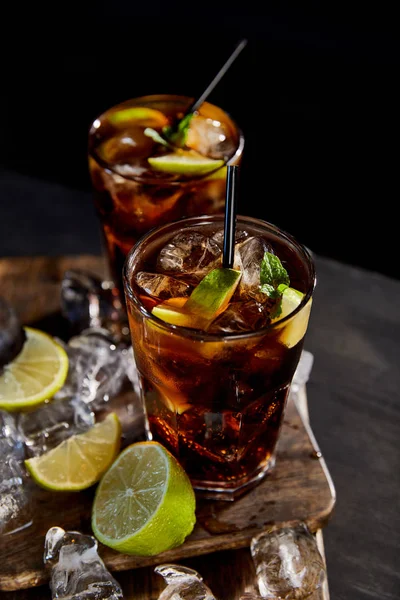 Vista de alto ángulo de cócteles cuba libre en vasos con pajitas, cubitos de hielo y limas sobre fondo negro - foto de stock