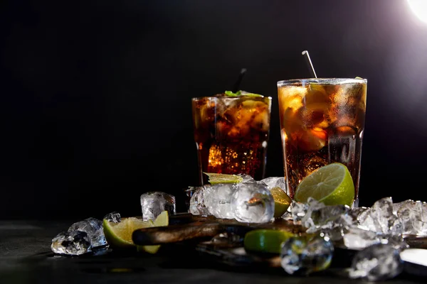 Cocktails cuba libre in Gläsern mit Strohhalmen, Eiswürfeln und Limetten auf schwarzem Hintergrund — Stockfoto