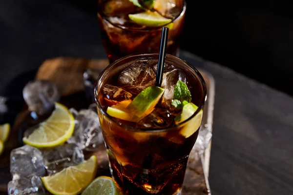Selektiver Fokus von Cocktails cuba libre in Gläsern mit Strohhalmen, Eiswürfeln und Limetten auf schwarzem Hintergrund — Stockfoto