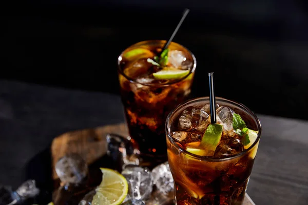 Cocktails cuba libre in glasses with straws, ice cubes and limes on black background — Stock Photo