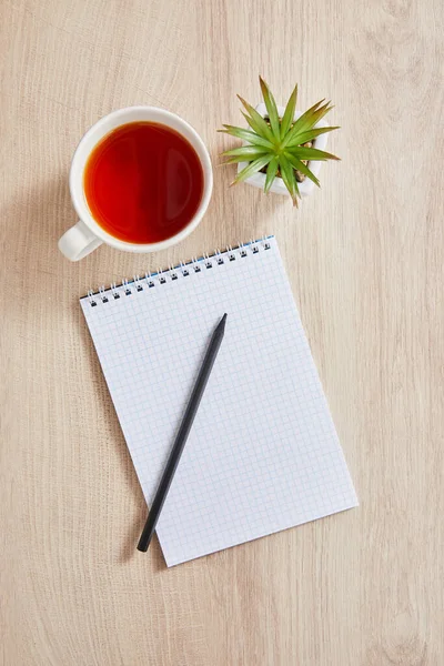 Top view of green plant, cup of tea and blank notebook with pencil on wooden surface — Stock Photo