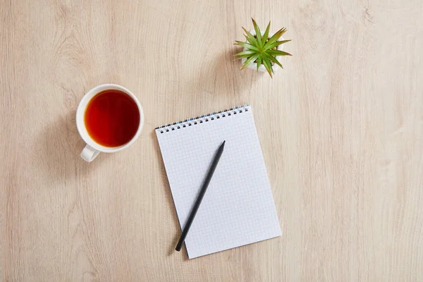 Vue de dessus de la plante verte, tasse de thé et bloc-notes vierge avec crayon sur la surface en bois — Photo de stock