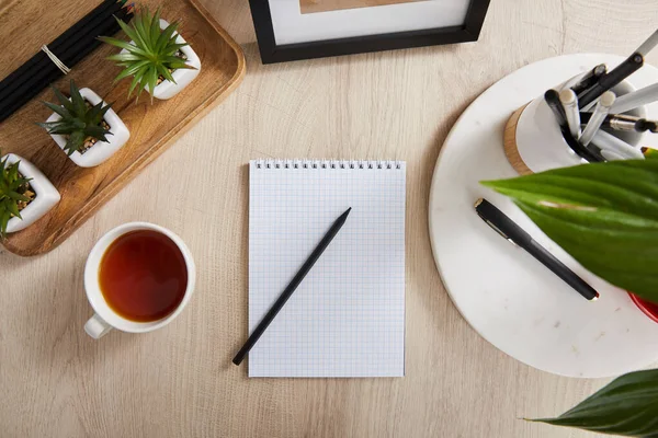 Vista superior de las plantas verdes, taza de té y cuaderno en blanco con lápices y bolígrafos en la superficie de madera - foto de stock