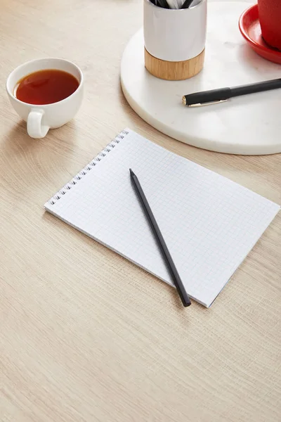 Taza de té y cuaderno en blanco con lápiz y pluma en la superficie de madera - foto de stock