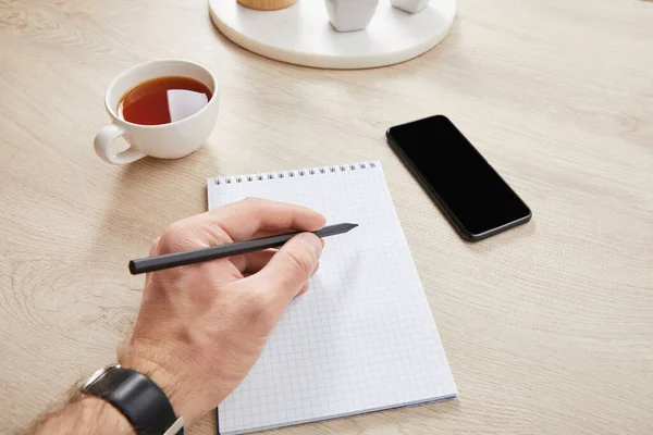 Cropped view of man writing in notebook near cup of tea and smartphone on wooden surface — Stock Photo