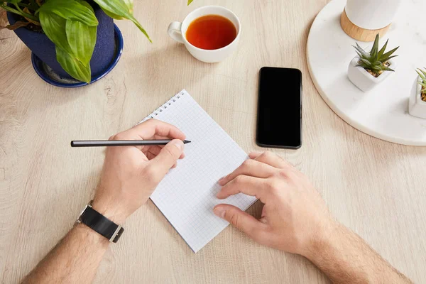 Cropped view of man writing in notebook near green plants, cup of tea and smartphone on wooden surface — Stock Photo