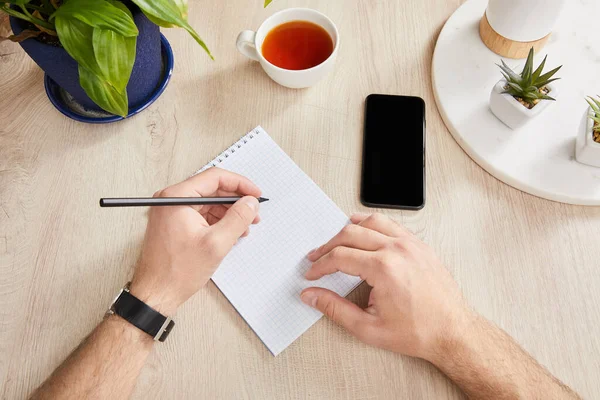 Vista recortada del hombre escribiendo en un cuaderno cerca de plantas verdes, taza de té y teléfono inteligente en la superficie de madera — Stock Photo