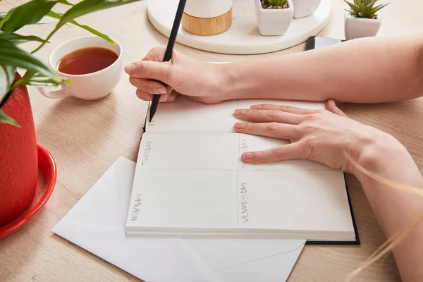 Cropped view of woman writing in notebook near green plants, cup of tea on wooden surface — Stock Photo