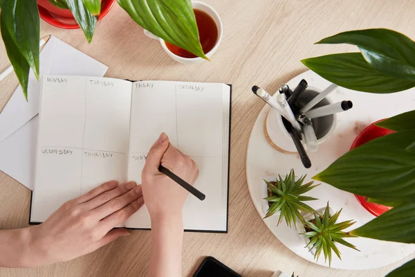 Cropped view of woman writing in notebook near green plants, cup of tea on wooden surface — Stock Photo