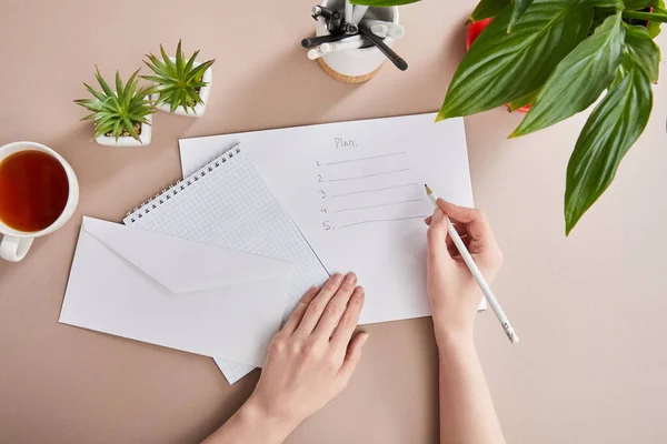 Обрезанный вид женщины, пишущей план на бумаге рядом с зелеными растениями, чашка чая, конверт, чистый блокнот на бежевой поверхности — стоковое фото