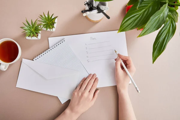 Vue recadrée du plan d'écriture femme sur papier près des plantes vertes, tasse de thé, enveloppe, carnet vierge sur surface beige — Photo de stock