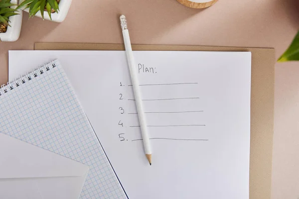 Draufsicht auf grüne Pflanzen, Umschlag, leeres Notizbuch, Bleistifte und Papier mit Planschrift auf beiger Oberfläche — Stockfoto