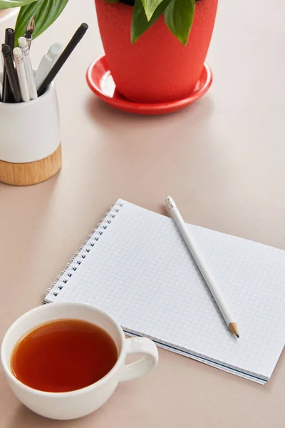 Green plant, cup of tea, blank notebook with pencils on beige surface — Stock Photo