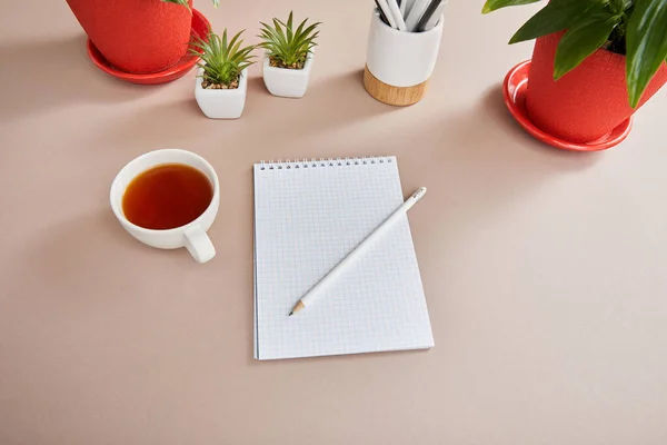 Green plants, cup of tea, blank notebook with pencil on beige surface — Stock Photo