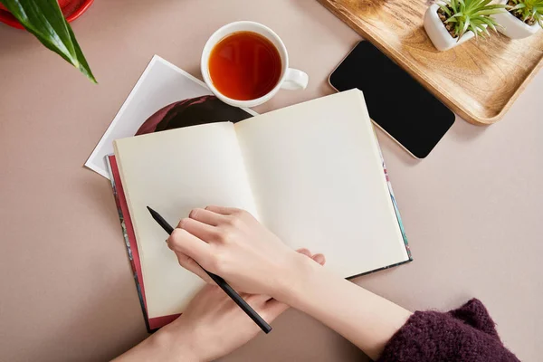 Ausgeschnittene Ansicht einer Frau, die in Notizbuch neben grünen Pflanzen auf Holztafel schreibt, Tasse Tee, Smartphone auf beiger Oberfläche — Stockfoto