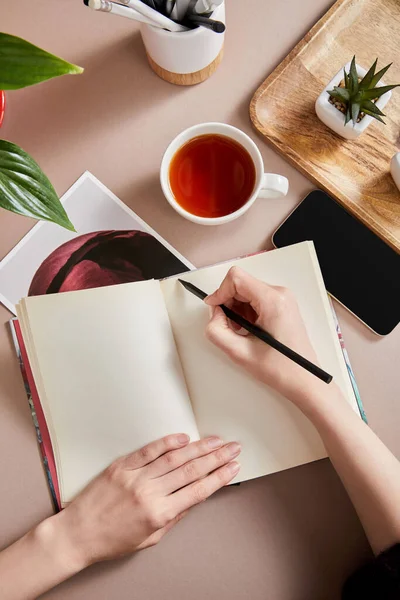 Vista recortada de la mujer escribiendo en planificador cerca de plantas verdes en tablero de madera, taza de té, teléfono inteligente en la superficie beige - foto de stock