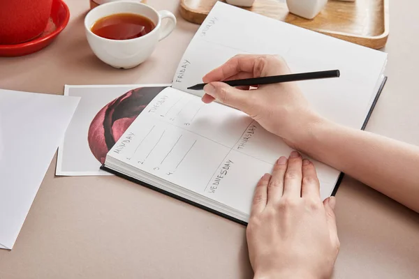 Cropped view of woman writing in planner near cup of tea on beige surface — Stock Photo