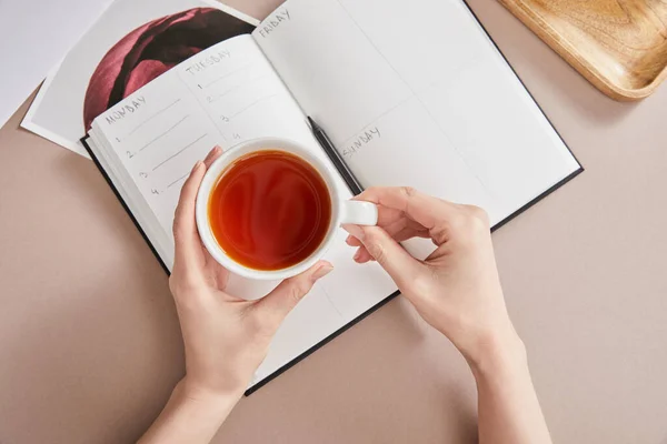 Верхний вид женских рук с чашкой чая возле планировщика с карандашом на бежевой поверхности — стоковое фото