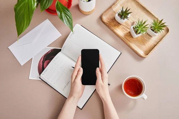 Ansicht weiblicher Hände mit Smartphone in der Nähe grüner Pflanzen auf Holzbrett, Tasse Tee, Planer mit Bleistift auf beiger Oberfläche — Stockfoto