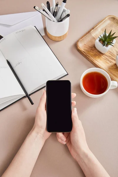 Frauenhände mit Smartphone in der Nähe grüner Pflanzen auf Holzbrett, Tasse Tee, Planer mit Bleistiften und Umschlag auf beiger Oberfläche — Stockfoto