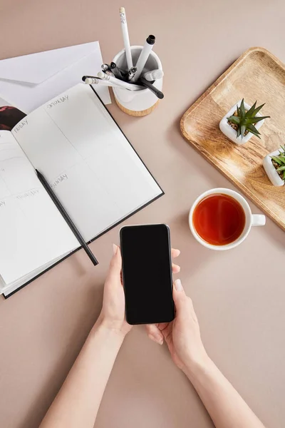 Vista superior de manos femeninas con teléfono inteligente cerca de plantas verdes en tablero de madera, taza de té, planificador con lápices y sobre en superficie beige - foto de stock