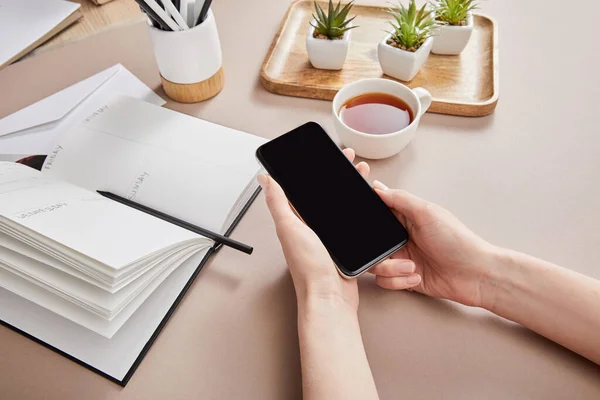 Female hands with smartphone near green plants on wooden board, cup of tea, planner with pencils and envelope on beige surface — Stock Photo