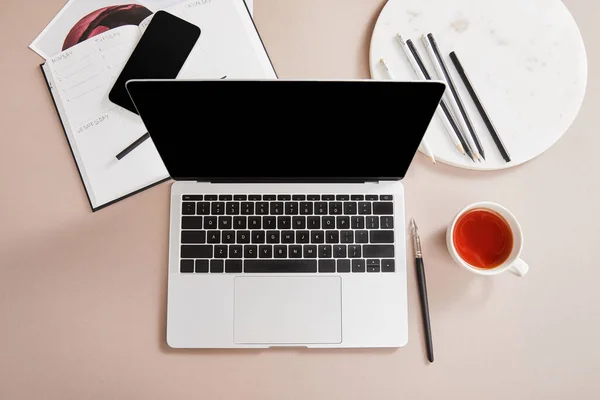 Top view of cup of tea, laptop, planner, card, smartphone near circle with pencils on beige surface — Stock Photo