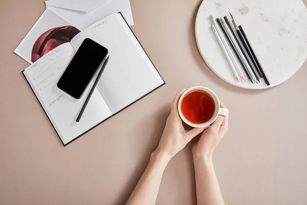 Cropped view of woman holding cup of tea near planner, card, smartphone near circle with pencils on beige surface — Stock Photo
