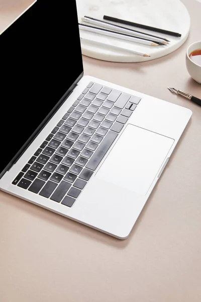 Ноутбук с чистым экраном возле чашки чая и канцелярских принадлежностей на бежевой поверхности — стоковое фото