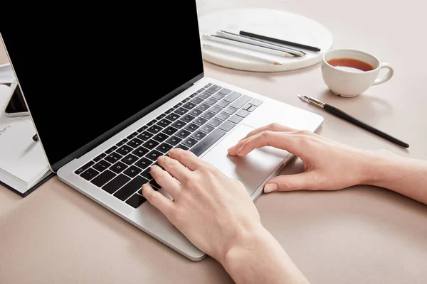 Cropped view of woman using laptop near cup of tea on beige surface — Stock Photo