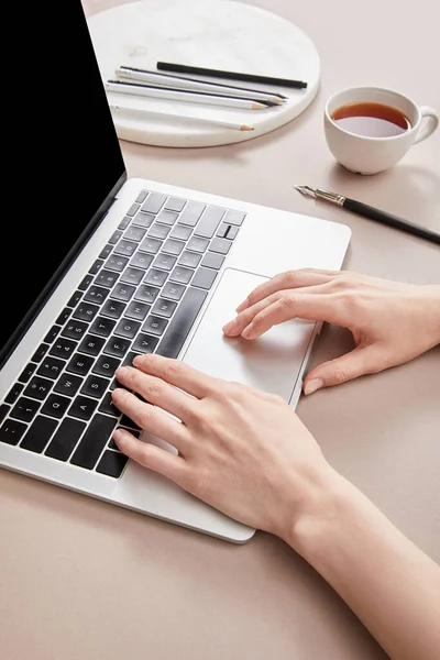 Vista recortada de la mujer usando el ordenador portátil cerca de la taza de té en la superficie beige - foto de stock