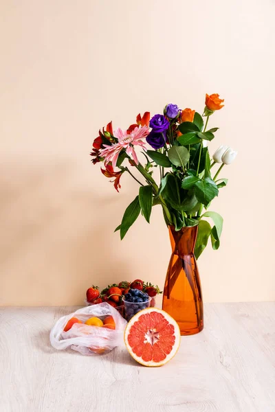 Floral and fruit composition with bouquet in vase, berries, grapefruit and apricots on wooden surface on beige background — Stock Photo