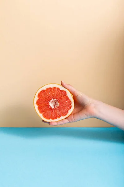 Vista recortada de la mano femenina con jugosa mitad pomelo sobre fondo beige y azul - foto de stock