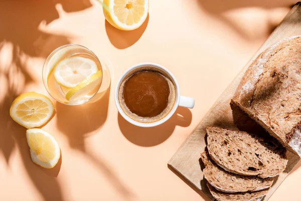 Blick von oben auf Kaffeetasse, Glas Wasser mit Zitrone und Brot zum Frühstück auf beigem Tisch — Stockfoto
