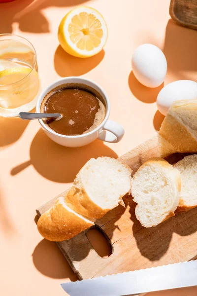 Coffee cup with teaspoon, eggs, baguette and glass of water with lemon for breakfast on beige table — Stock Photo