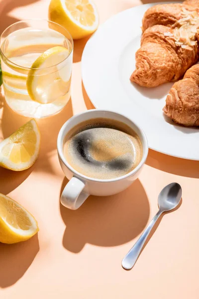 Kaffeetasse mit Teelöffel, Croissants und Glas Wasser mit Zitrone zum Frühstück auf beigem Tisch — Stockfoto