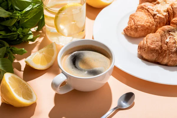 Kaffee, Croissants und ein Glas Wasser mit Zitrone zum Frühstück auf einem beigen Tisch — Stockfoto