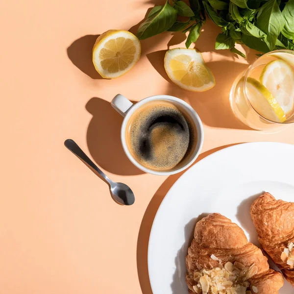 Верхний вид на чашку кофе, зелени, лимоны и круассаны на завтрак на бежевый стол — стоковое фото