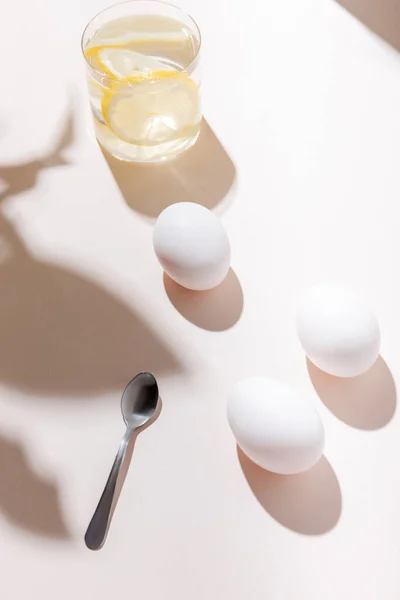 Chicken eggs, teaspoon and glass of water with lemon for breakfast on grey table with shadows — Stock Photo