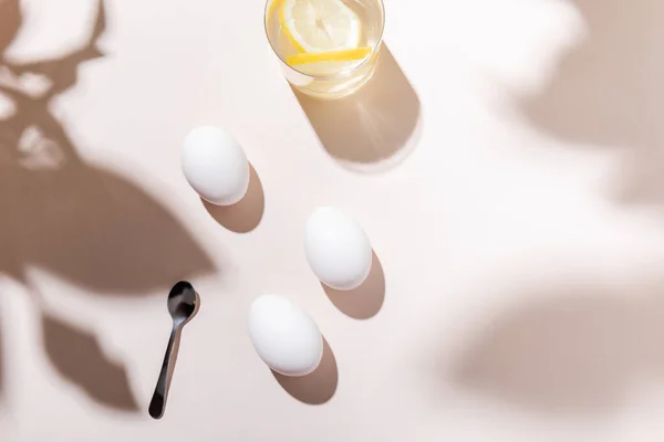 Top view of white boiled chicken eggs, water with lemon and teaspoon on grey table with shadows — Stock Photo