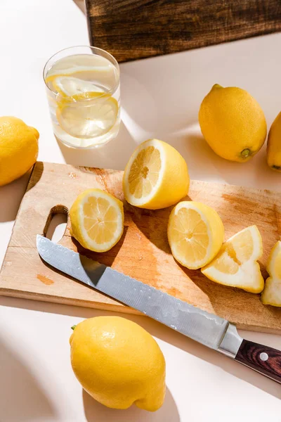 Whole and cutted lemons on wooden board with knife and glass of water on grey table — Stock Photo