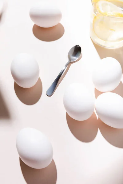Белые куриные яйца, чайная ложка и стакан воды с лимоном на завтрак на сером столе с тенями — стоковое фото