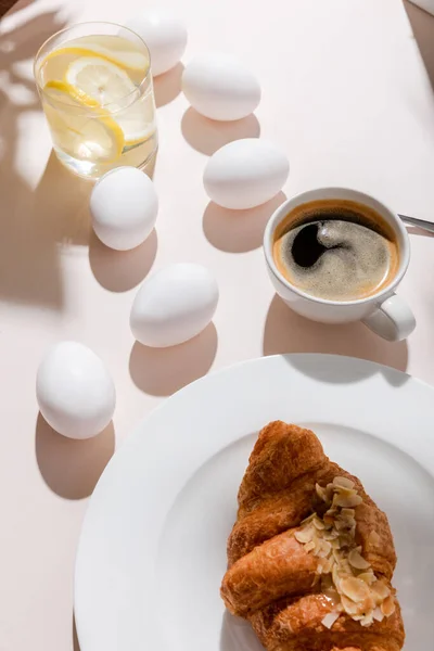 Uova di pollo bollite biologiche, croissant, tazza di caffè e bicchiere d'acqua con limone a colazione su tavolo grigio con ombre — Foto stock