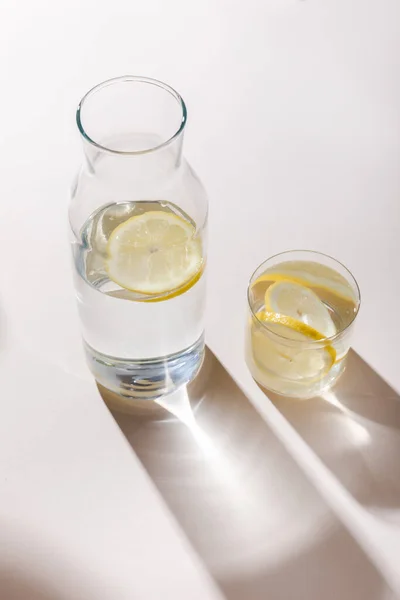 Agua dulce con rodajas de limón en jarra y vidrio sobre mesa blanca con sombras - foto de stock