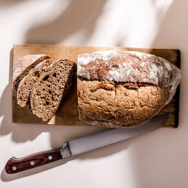 Vista superior de pão fresco assado e fatiado a bordo com faca na mesa cinza com sombras — Fotografia de Stock