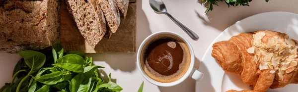 Vista superior de cruasanes frescos, pan, albahaca y taza de café para el desayuno en la mesa gris, encabezado del sitio web - foto de stock