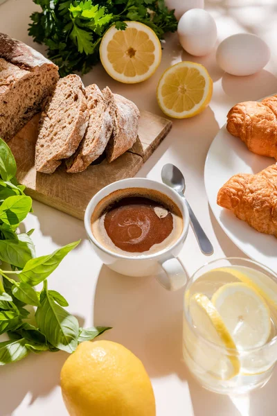 Fresh croissants, bread, greenery, eggs, lemon water and cup of coffee for breakfast on grey table — Stock Photo