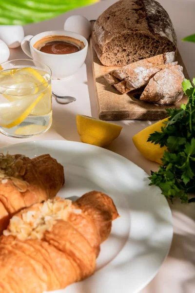 Cruasanes frescos, pan, huevos, café y agua de limón para el desayuno en la mesa gris, enfoque selectivo - foto de stock